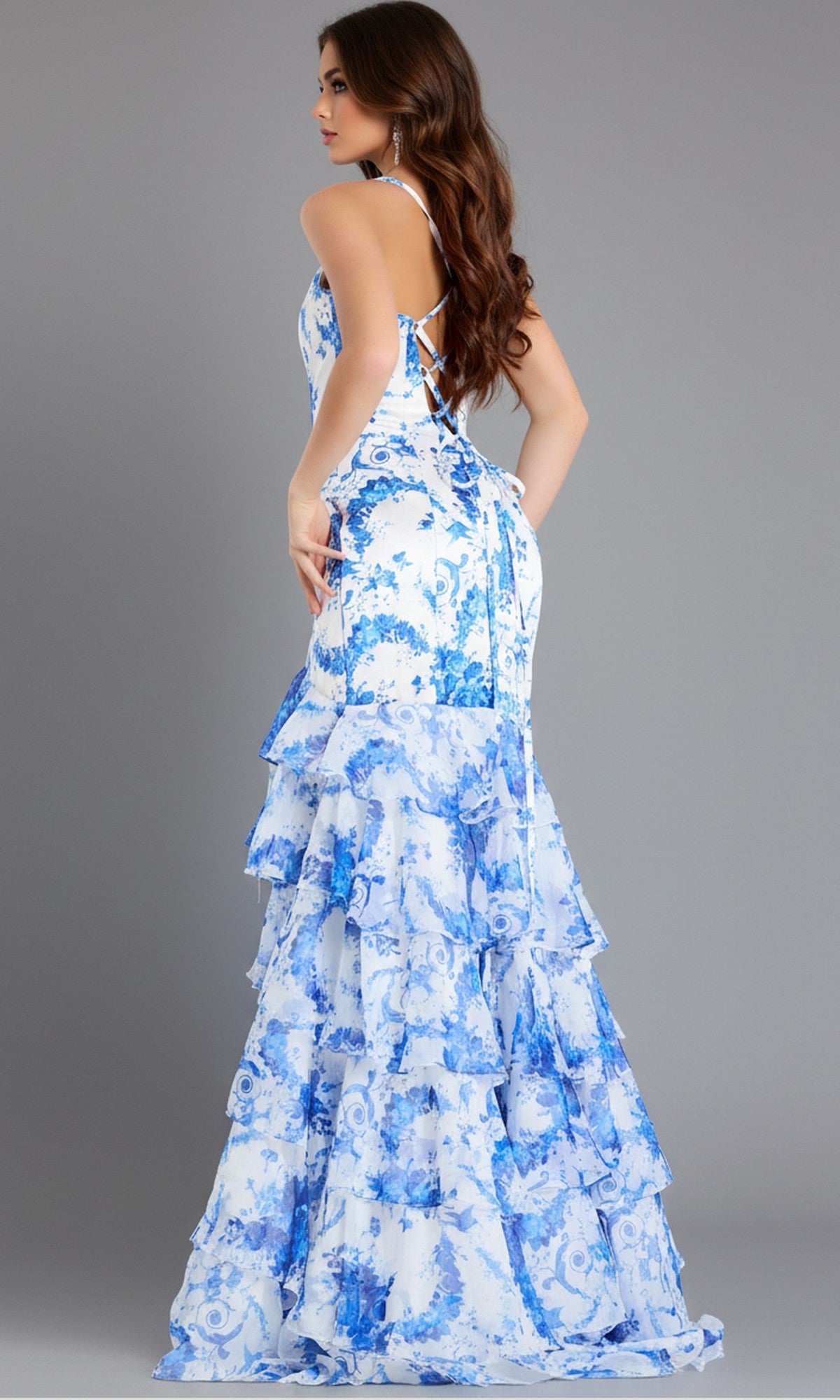 A woman poses in the Jovani 48331 long prom dress, which features a floral print, lace-up open back, and tiered white and blue ruffle skirt, set against a plain gray background.